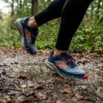 Gros plan sur un coureur marchant dans une flaque d'eau en forêt avec des chaussures de running bleu et orange.