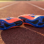 Une paire de chaussures de sprint orange et bleue avec des pointes, posée sur une piste d'athlétisme en tartan.