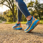 Gros plan sur des chaussures de course bleues et orange portées par un coureur sur un sentier en terre.