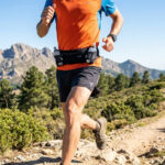 Un coureur de trail portant une ceinture porte-bidon sur un sentier de montagne ensoleillé.