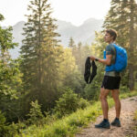 Un coureur en forêt hésite entre un sac d'hydratation bleu et une ceinture de running noire lors d'une sortie en montagne.