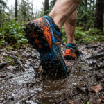 Gros plan sur une chaussure de trail bleue et orange courant dans une flaque de boue en forêt.