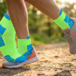 Coureur portant des chaussettes de sport néon vert et bleu sur un sentier en forêt lors d'une journée ensoleillée.