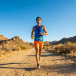 Un coureur en tenue de sport porte une gourde en courant sur un sentier en plein désert sous un ciel bleu dégagé.