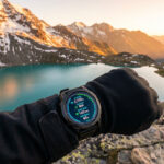 Une montre Garmin Fenix 8 au poignet affichant des données biométriques devant un lac de montagne au coucher du soleil.
