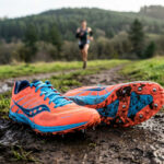 Paire de chaussures d'athlétisme orange et bleue posée sur un chemin boueux, avec un coureur en arrière-plan.