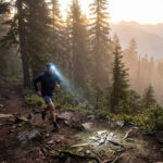 Un coureur de trail équipé d'une lampe frontale court sur un sentier en forêt au lever du soleil.