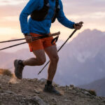 Un coureur de trail portant un sac à dos et tenant des bâtons pliables sur un sentier montagneux au coucher du soleil.