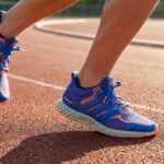 Un coureur porte des baskets Adidas bleues à semelles imprimées en 3D sur une piste d'athlétisme au coucher du soleil.