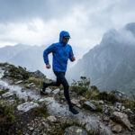 Un coureur de trail portant une veste bleue imperméable court sous la pluie sur un sentier montagneux escarpé.